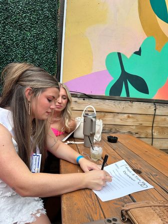 Two women at an outdoor patio wooden table — one in a white dress filling out a "Bachelorette Experience" form, colorful mural and faux greenery backdrop, drinks and handbags nearby.