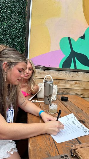 Two women at an outdoor patio wooden table — one in a white dress filling out a "Bachelorette Experience" form, colorful mural and faux greenery backdrop, drinks and handbags nearby.