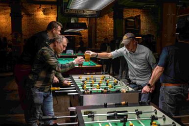 Men clinking beer glasses over a foosball table in a dim, brick-walled pub with pool tables in the background — casual bar game night.