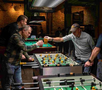 Men clinking beer glasses over a foosball table in a dim, brick-walled pub with pool tables in the background — casual bar game night.