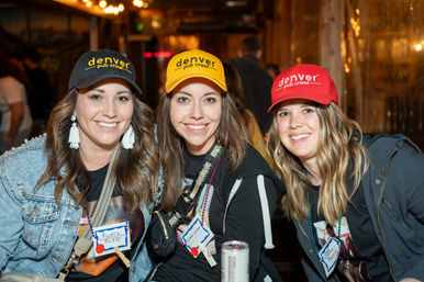 Three smiling women wearing colorful Denver caps and name tags at a lively Denver bar during a pub crawl, posing together with drinks