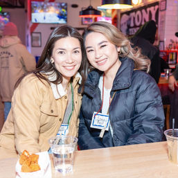 Two smiling women in winter jackets at a lively neighborhood bar counter with drinks, a small snack basket, and colorful lights