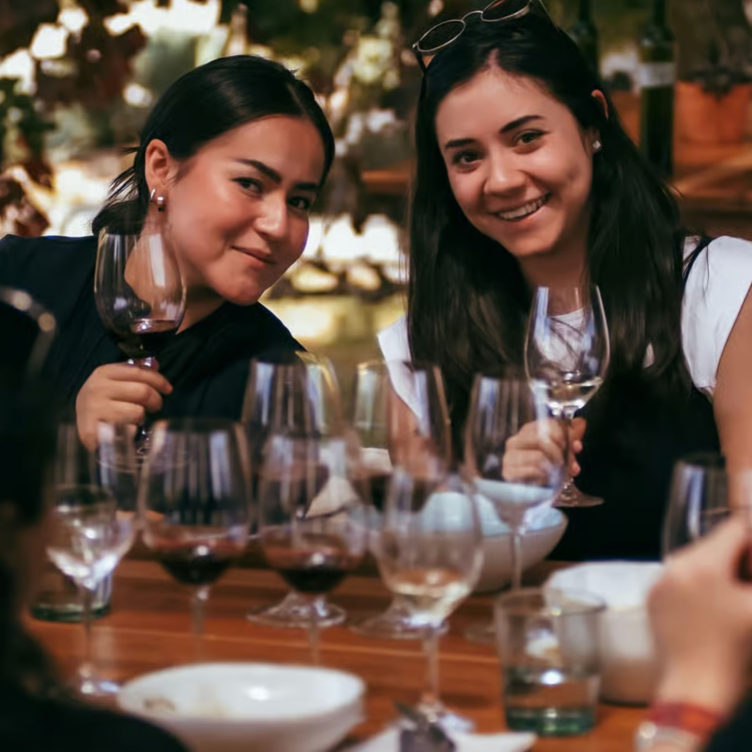 Two friends smiling at an outdoor wine tasting, holding wine glasses at a wooden table with multiple poured glasses and bowls.