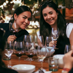 Two friends smiling at an outdoor wine tasting, holding wine glasses at a wooden table with multiple poured glasses and bowls.