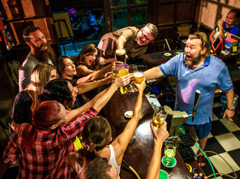 Lively group at a neighborhood pub gathered around a wooden bar, raising beer glasses for a joyful toast with a smiling bartender during a festive night out.
