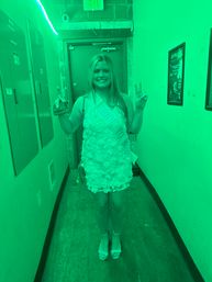 Smiling woman in a floral textured mini dress wearing a celebratory sash, flashing peace signs in a narrow indoor hallway bathed in green light