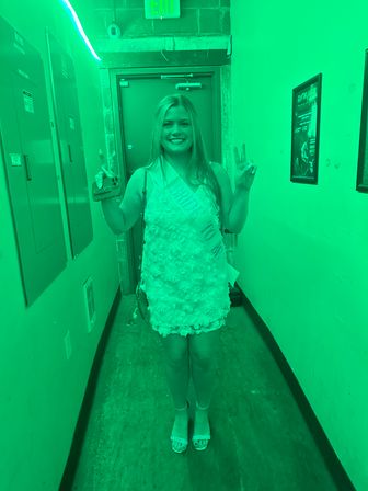 Smiling woman in a floral textured mini dress wearing a celebratory sash, flashing peace signs in a narrow indoor hallway bathed in green light