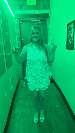 Smiling woman in a floral textured mini dress wearing a celebratory sash, flashing peace signs in a narrow indoor hallway bathed in green light