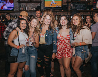 Five smiling women in summer outfits — denim skirts, ripped jeans, and a red floral romper — holding drinks and posing for a group photo inside a lively sports bar with TVs and exposed brick.