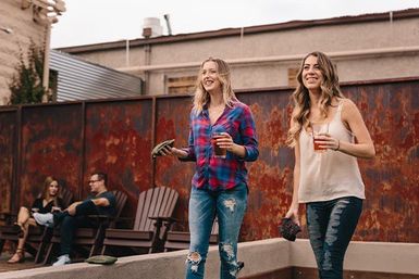 Two friends playing cornhole on a rustic urban rooftop patio, holding drinks and tossing bean bags while others relax in Adirondack chairs by a weathered metal fence.