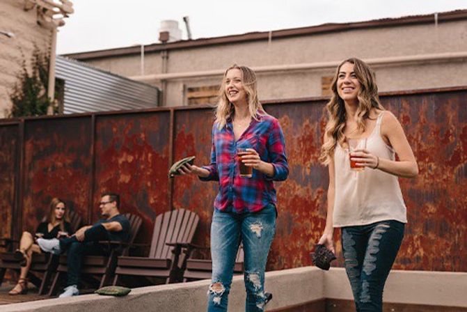 Two friends playing cornhole on a rustic urban rooftop patio, holding drinks and tossing bean bags while others relax in Adirondack chairs by a weathered metal fence.