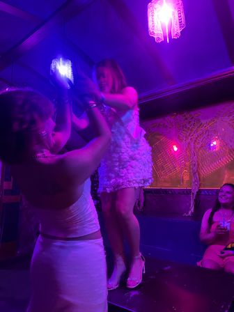 Friends dancing on a raised platform under purple neon lights in a lively bar/nightclub, woman in a white floral dress and heels helped by a friend while another guest smiles with a drink.
