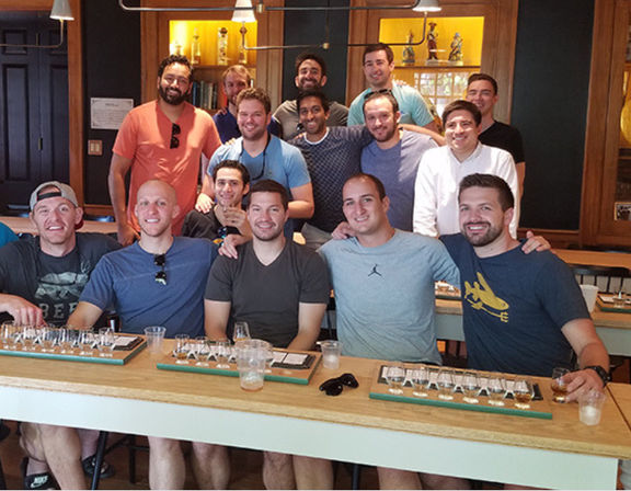 Smiling group of men posing in a warm, wood-paneled tasting room with whiskey flight glasses and small pours lined up on a long bar.