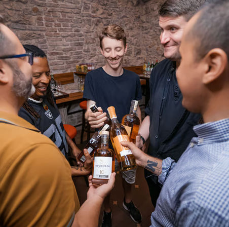 Group of five people clinking whiskey bottles in a cozy brick‑walled bar during a casual whiskey tasting night out.