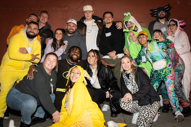 Nighttime group photo of adults in colorful costumes and casual clothes posing and smiling against a red urban wall, featuring onesies, animal and character outfits and visible name tags.