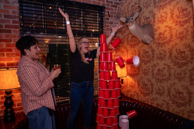 Two people in a cozy brick-walled bar cheer as a tall tower of red plastic cups collapses near a mounted deer head and vintage deer-print wallpaper, warm lamps and string lights adding nightlife, party vibe.