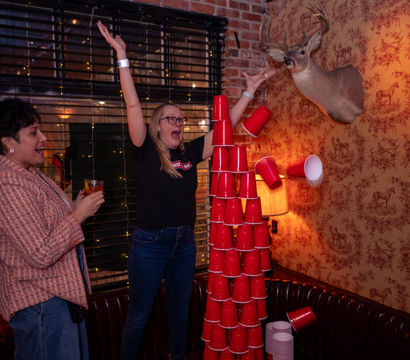 Two people in a cozy brick-walled bar cheer as a tall tower of red plastic cups collapses near a mounted deer head and vintage deer-print wallpaper, warm lamps and string lights adding nightlife, party vibe.