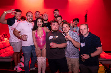 Group of friends posing in a red-lit nightclub lounge — woman in a shiny pink bodysuit and white thigh-high boots at center, men around her holding drinks and smiling in a casual party scene.