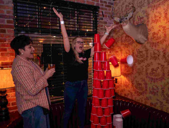 Two guests cheer as a tall pyramid of red plastic cups topples during a party in a cozy brick-walled bar with deer-motif wallpaper and a mounted deer head