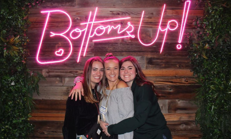 Three smiling women hugging and posing in front of a pink neon "Bottoms Up!" sign on a wood-paneled wall with green plants — fun nightlife photo backdrop.