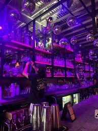 Neon-purple lit bar interior with hanging disco balls, mirrored shelves stocked with liquor and glassware, and a bartender reaching for bottles while prepping drinks — vibrant nightlife scene.