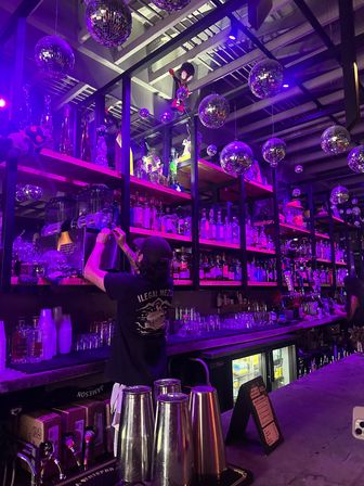 Neon-purple lit bar interior with hanging disco balls, mirrored shelves stocked with liquor and glassware, and a bartender reaching for bottles while prepping drinks — vibrant nightlife scene.