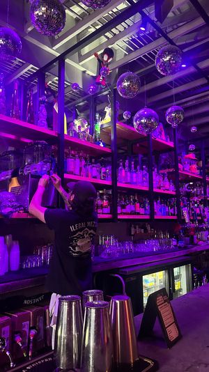 Neon-purple lit bar interior with hanging disco balls, mirrored shelves stocked with liquor and glassware, and a bartender reaching for bottles while prepping drinks — vibrant nightlife scene.