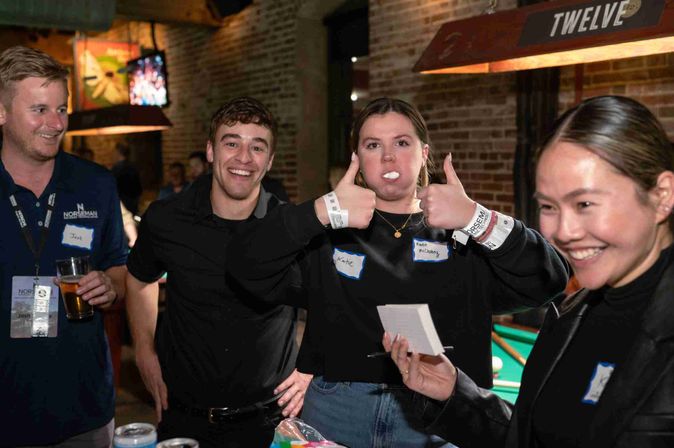 Smiling group of friends at a brick-walled bar with a pool table, playful woman giving double thumbs-up with a white object in her mouth.