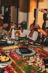 Cheerful group toasting at an indoor dinner party around a tropical-themed table decorated with palm leaves, rose petals, tacos and appetizers