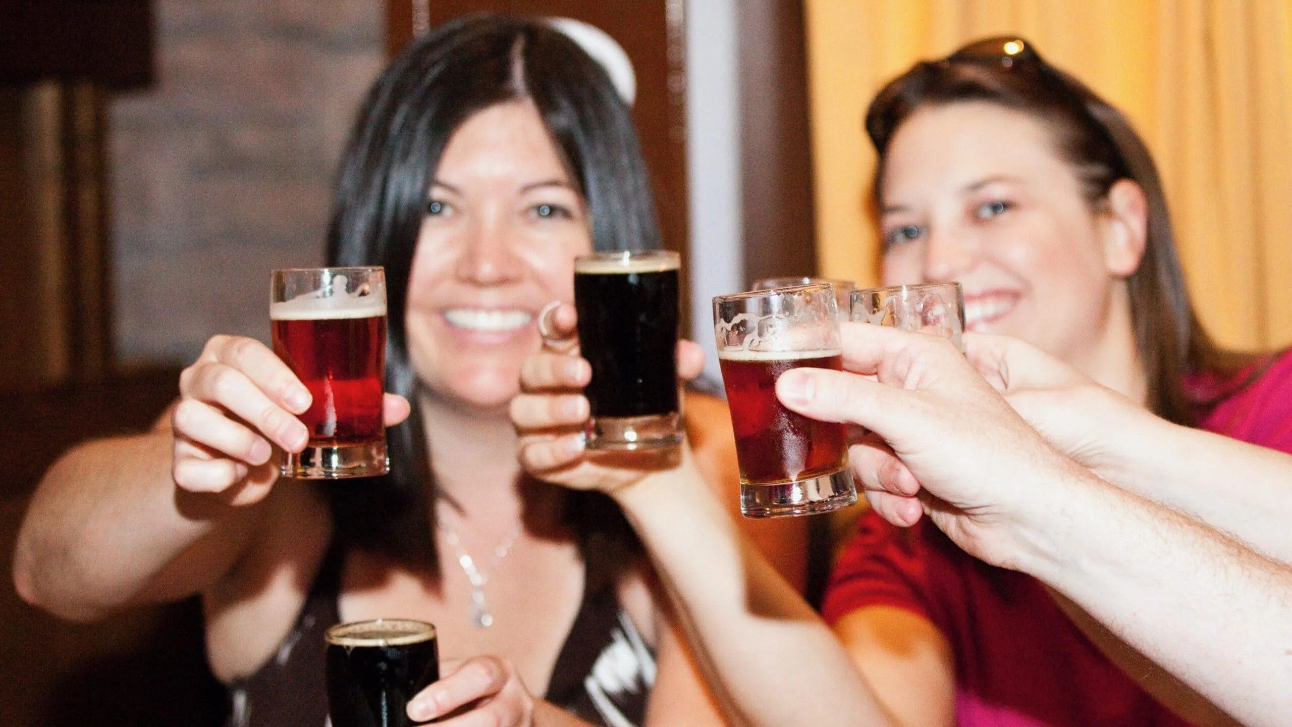 Friends cheer with small craft beer flight glasses in a cozy brewery tasting room, smiling and toasting