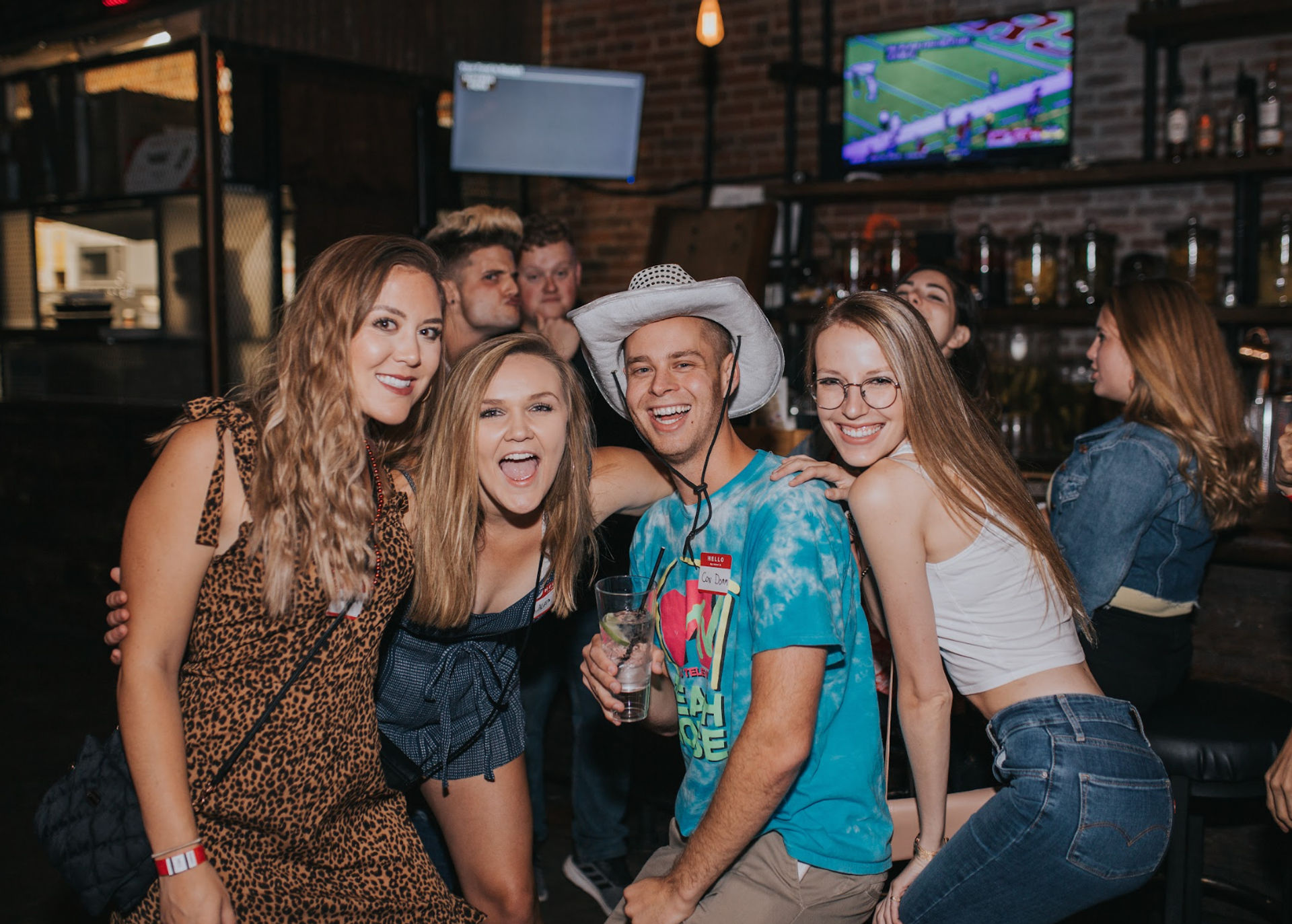 Group of friends laughing and posing at a lively downtown bar at night — a man in a white cowboy hat holding a cocktail and three women in casual outfits with TVs and brick bar shelving behind them.