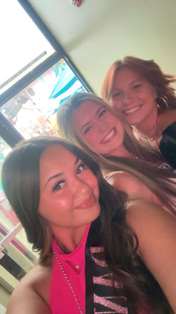 Three smiling friends taking a close-up selfie at a colorful indoor nightlife venue, wearing party beads and a glittery sash with bright neon signage visible through a window behind them.