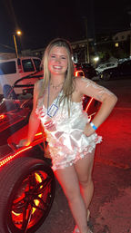 Smiling woman in a white floral romper and sash posing by a red LED-lit three-wheeled vehicle at night in a parking lot, festive party vibe.