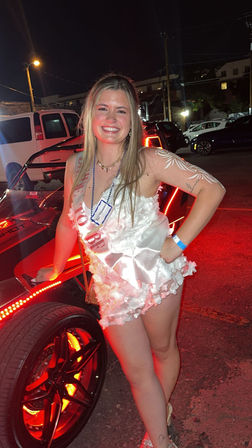 Smiling woman in a white floral romper and sash posing by a red LED-lit three-wheeled vehicle at night in a parking lot, festive party vibe.