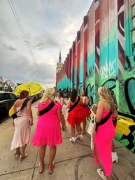 Group of women in bright pink dresses and black sashes walking along a city sidewalk beside a colorful graffiti mural and distant church steeple — playful bachelorette-style outing with a yellow umbrella.