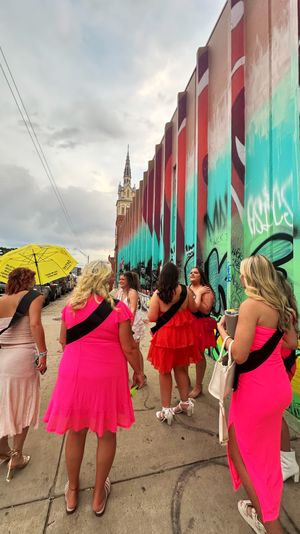 Group of women in bright pink dresses and black sashes walking along a city sidewalk beside a colorful graffiti mural and distant church steeple — playful bachelorette-style outing with a yellow umbrella.