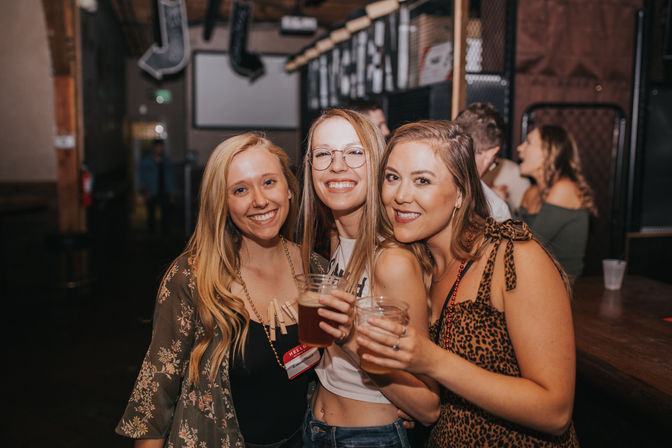 Three smiling friends holding plastic cups of beer and posing together at an indoor bar for a casual night out with warm ambient lighting.