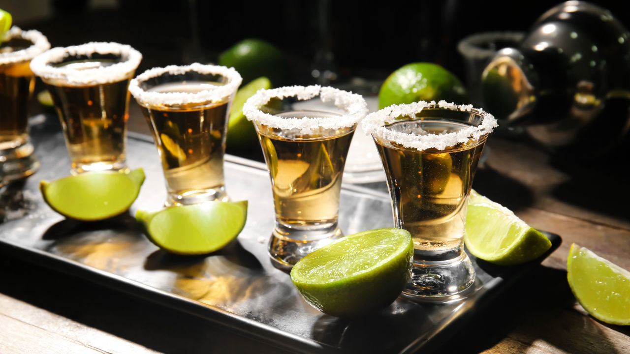 Golden tequila shots in salt-rimmed glasses with bright lime wedges arranged on a metal tray at a bar