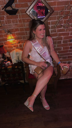 Smiling guest in a white dress wearing a "To Be" sash seated on a wooden chair against a brick wall, holding a cocktail with crossed legs in sparkly heels, small purse and phone on her lap, warm lamp and mirror decor suggesting a bachelorette night out