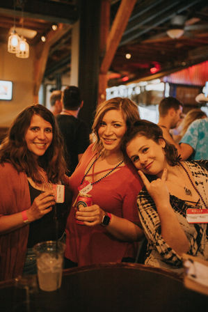 Three women smiling at a cozy wood‑beamed bar, holding drinks and wearing name tags and bead necklaces — lively nightlife scene.