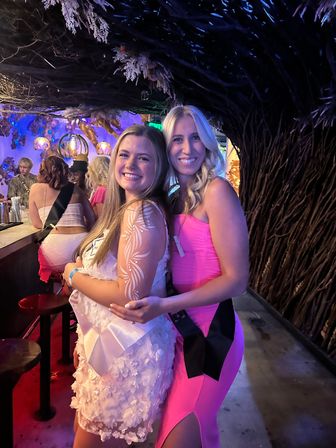 Two smiling women in pink party dresses posing at a dimly lit bar with woven branch wall decor, colorful hanging lights, and a festive bachelorette-party vibe.