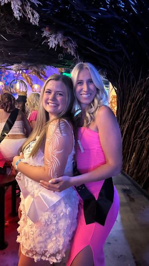 Two smiling women in pink party dresses posing at a dimly lit bar with woven branch wall decor, colorful hanging lights, and a festive bachelorette-party vibe.