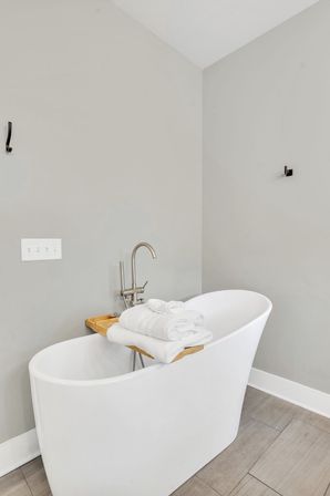 Minimalist modern bathroom interior with white freestanding bathtub, wooden bath tray holding folded white towels, nickel faucet, neutral gray walls and wood-look tile floor.