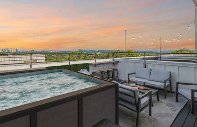 Bubbly rooftop hot tub and modern outdoor lounge with string lights overlooking trees and a distant city skyline at a peach-pink sunset.