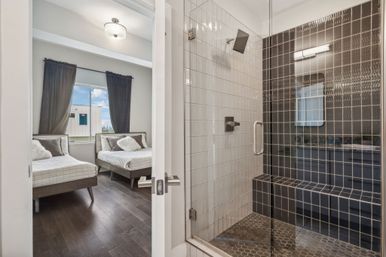 Sleek modern glass-enclosed shower with dark tiled bench and hexagon floor tiles opening to a cozy twin bedroom with dark hardwood floors, gray curtains and a distant city view in a contemporary apartment