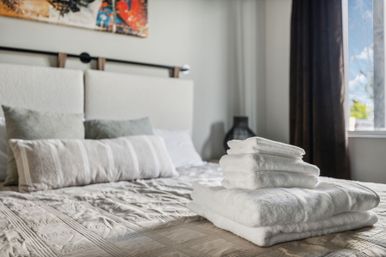 Fluffy white towels neatly stacked on a made bed in a sunlit modern bedroom with upholstered headboard, decorative pillows and a curtained window.