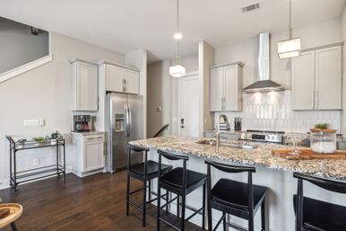 Modern open-concept kitchen with granite-topped island, four black bar stools, pendant lights, white cabinets, stainless steel refrigerator and range, and dark hardwood floors.