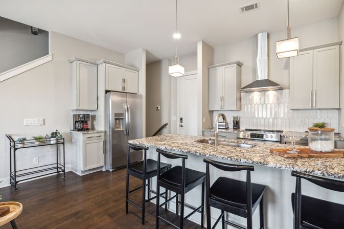 Modern open-concept kitchen with granite-topped island, four black bar stools, pendant lights, white cabinets, stainless steel refrigerator and range, and dark hardwood floors.