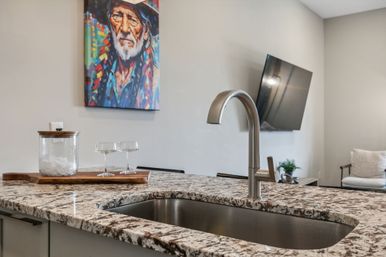 Modern open-plan kitchen island with speckled granite countertop, undermount stainless sink and curved faucet, glassware on a wooden tray and a vibrant portrait painting on the wall