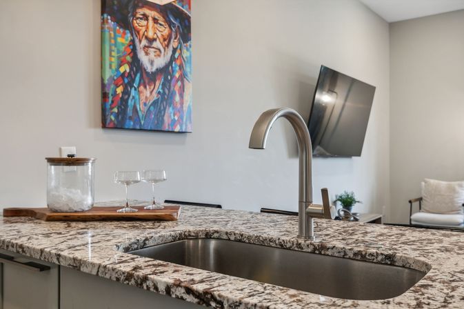 Modern open-plan kitchen island with speckled granite countertop, undermount stainless sink and curved faucet, glassware on a wooden tray and a vibrant portrait painting on the wall